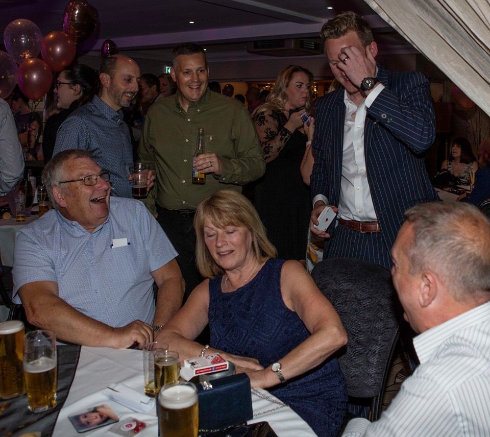 Party guests laughing around a table as Richard Kettle covers his face in mock disbelief