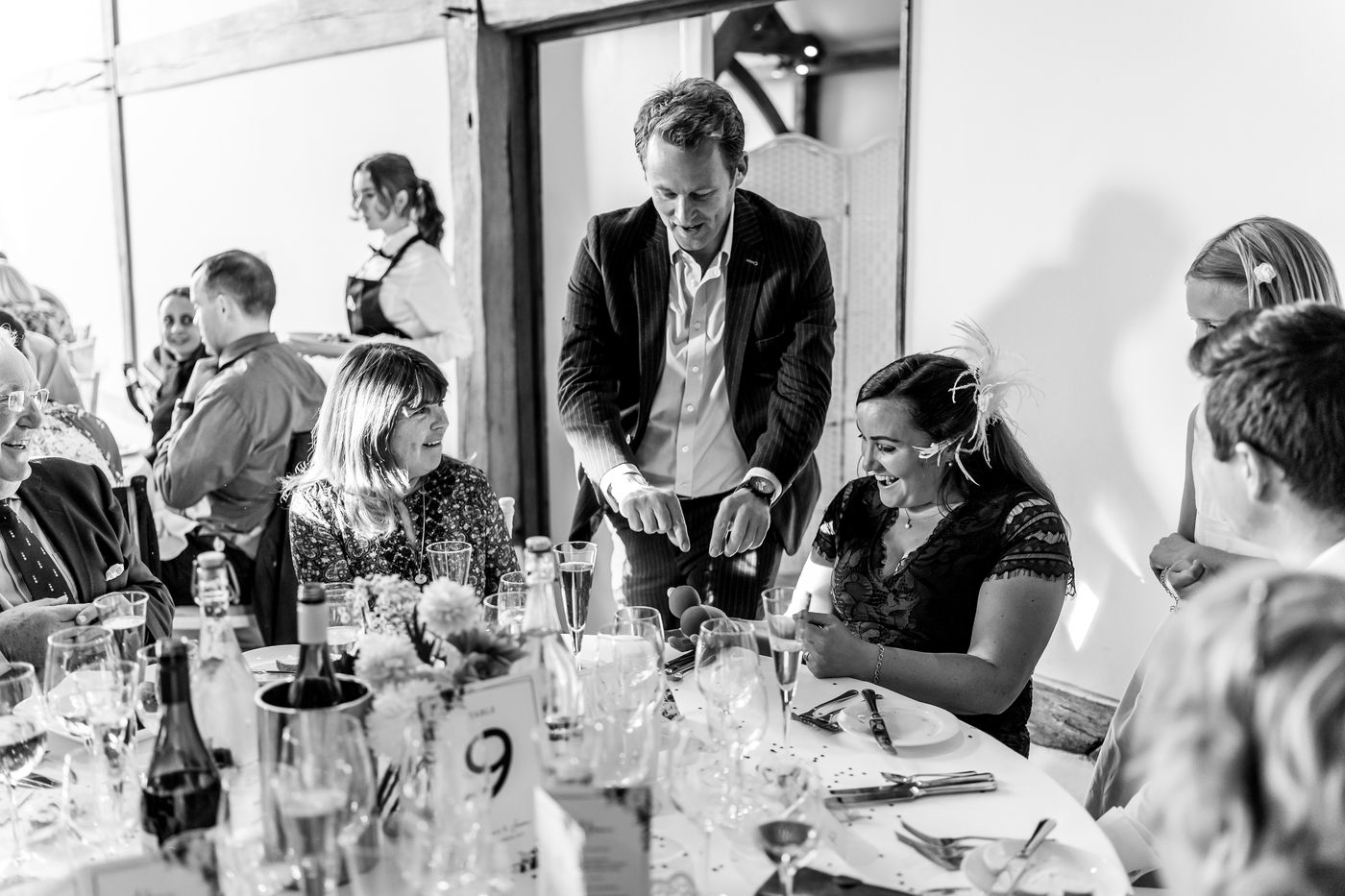 Wedding guests laughing during close-up magic at a barn reception, black and white