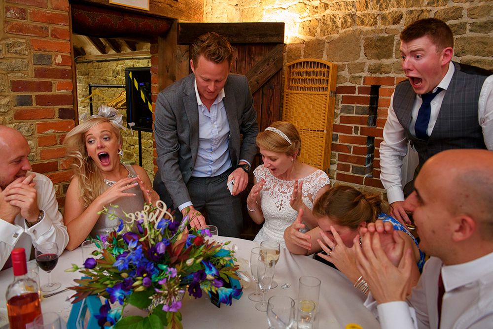 Wedding guests gasping and hiding their faces as a card trick lands at a stone barn reception